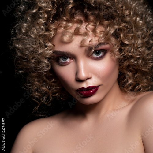 Cute caucasian woman with afro (curls) hairstyle on a dark background. She wears dark evening (or podium) make up with red mascara on her eyes ans lips