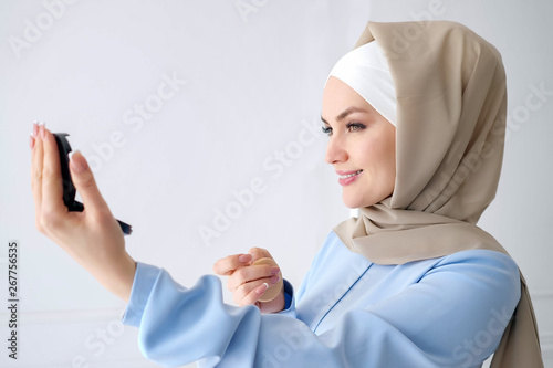 Portrait of young muslim woman in beige hijab and traditional blue dress looking at small mirror at home, side view.