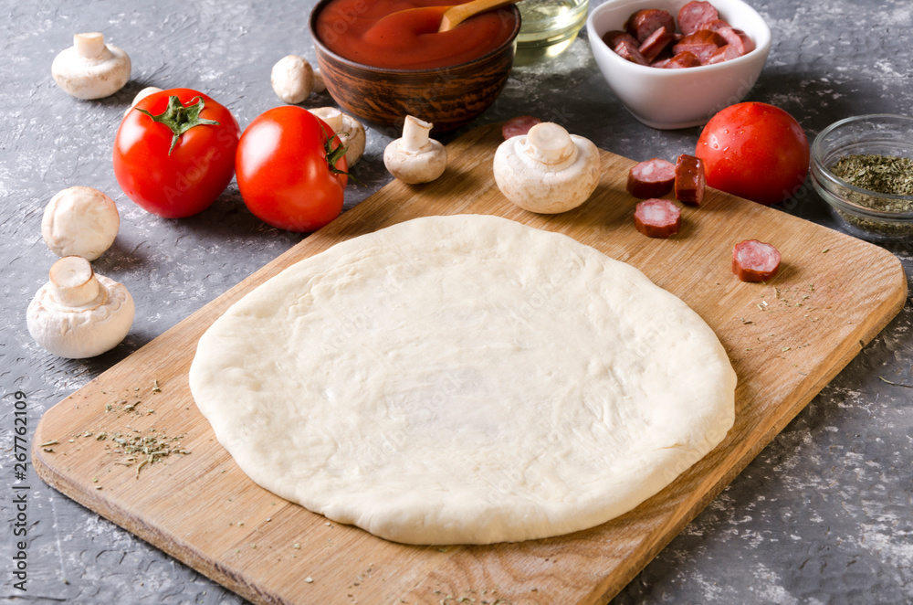 Closeup of raw dough prepared for pizza on the wooden board and different classic ingredients for it on the grey kitchen table Closeup of raw dough prepared for pizza on the wooden board and different classic ingredients for it on the grey kitchen table