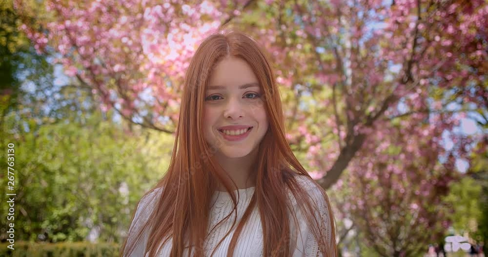 Attractive ginger female student in white blouse posing charmingly into camera in pink floral garden. Attractive ginger female student in white blouse posing charmingly into camera in pink floral garden.