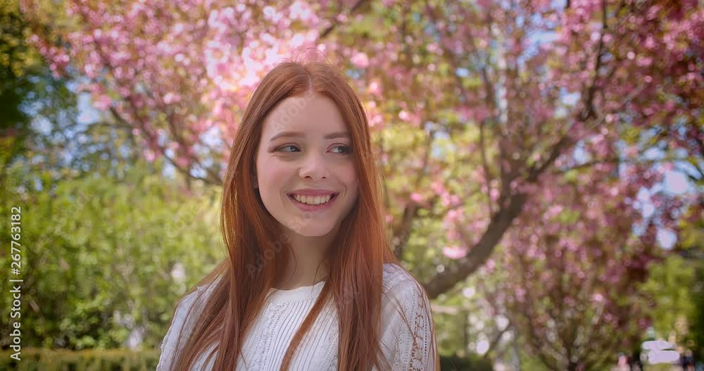 Charming ginger female student in white blouse posing gracefully into camera in pink floral garden. Charming ginger female student in white blouse posing gracefully into camera in pink floral garden.