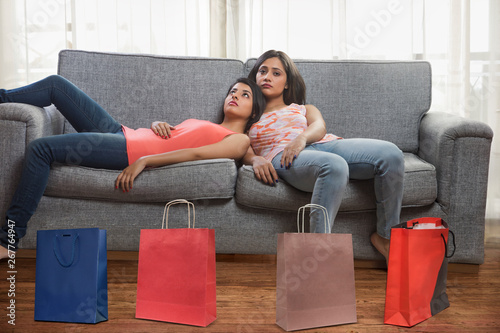 Two teenage girls lying exhausted on sofa at home after shopping with shopping bags on the floor	 photo