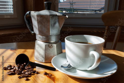 Coffee in a white cup and saucer with silver spoon, a tin of coffee beans, coffee beans and a coffee maker on a kitchen table in bright sunlight