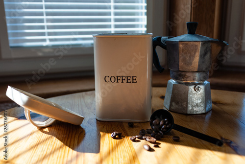 Coffee in a white cup and saucer with silver spoon, a tin of coffee beans, coffee beans and a coffee maker on a kitchen table in bright sunlight