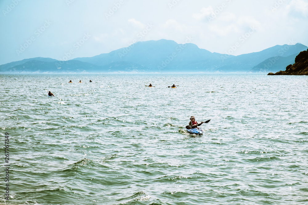 A group of unrecognizable people in kayaks moving on sea water near Hong Kong coastline. Island mountains in haze on background. Weekend activities. Extreme hobby and leisure of active men and women. A group of unrecognizable people in kayaks moving on sea water near Hong Kong coastline. Island mountains in haze on background. Weekend activities. Extreme hobby and leisure of active men and women.