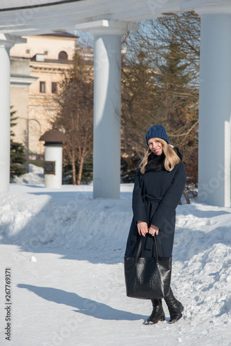 Young blonde girl in blue coat with fur in a knitted cap with a black big bag walks through the winter and spring city
