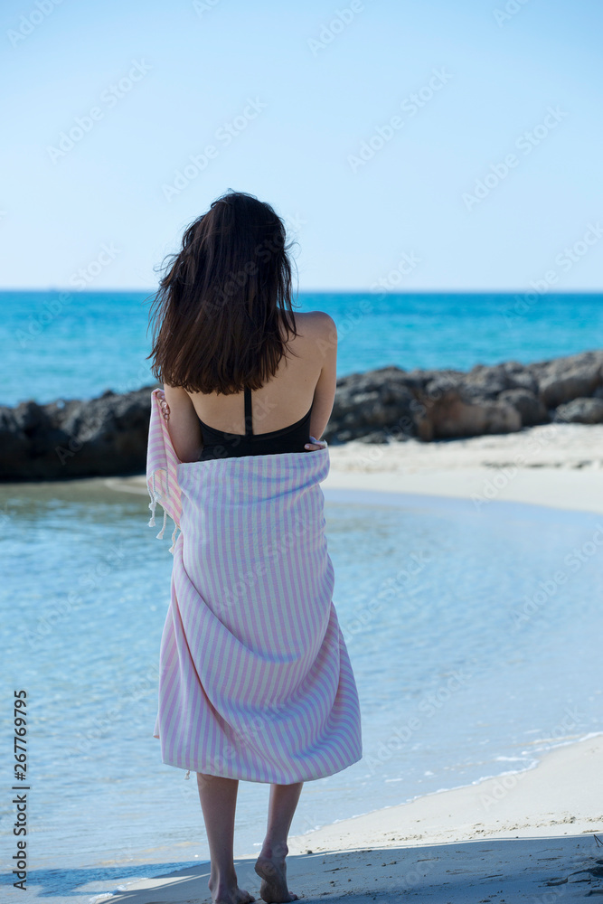 Young girl is on the beach and standing, posing to the camera. Beautiful girl, wrapped in pink white striped beach towel, sunny and windy weather. Background of blue sky, sea and beach - Image Young girl is on the beach and standing, posing to the camera. Beautiful girl, wrapped in pink white striped beach towel, sunny and windy weather. Background of blue sky, sea and beach - Image