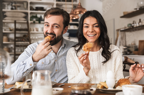 Image of young couple eating together at table while having breakfast in kitchen at home