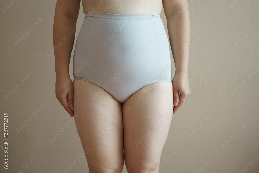 Curly cheerful overweight woman in white underwear stand near bed in  bedroom and put on slimming