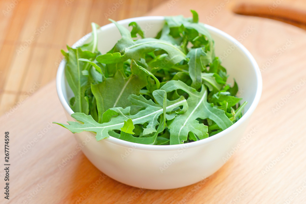 Fresh healthy green arugula leaves in a white bowl on light wooden background. Fresh healthy green arugula leaves in a white bowl on light wooden background.