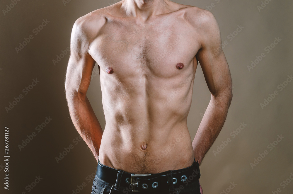 Fototapeta premium Naked torso of a young, slim, pumped-up man on a beige background. Tight naked attractive man in black jeans.