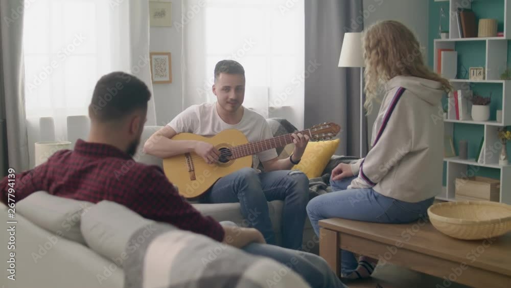 Handheld shot of young man plays guitar for his friends in a cozy living room. Handheld shot of young man plays guitar for his friends in a cozy living room.