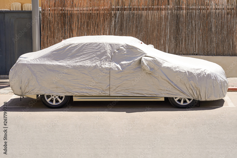 A car parked with protective cover silver. Car under a protective cover parked in the courtyard in sun weather, summer. Car with a protective, parking cover. The car is covered with a cover