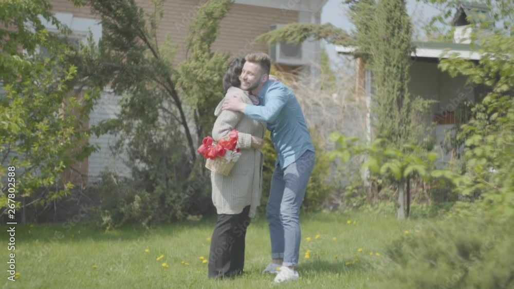 Old woman walking with the basket with tulips in it on the backyard. Adult grandson comes and hugs the lady, the guy visiting his granny. Family meeting, friendly relationships Old woman walking with the basket with tulips in it on the backyard. Adult grandson comes and hugs the lady, the guy visiting his granny. Family meeting, friendly relationships