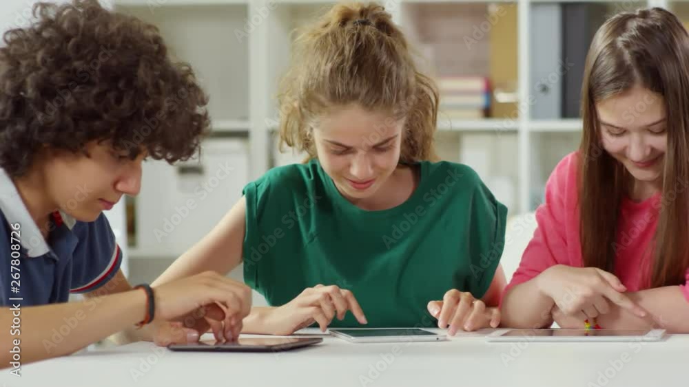 Tracking right of four Caucasian classmates sitting together at table, using tablets during break Tracking right of four Caucasian classmates sitting together at table, using tablets during break