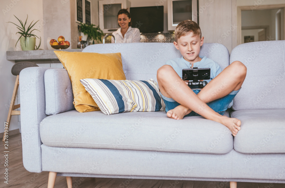 Boy plays with electronic device. His mother prepared something in the blurred background kitchen Boy plays with electronic device. His mother prepared something in the blurred background kitchen
