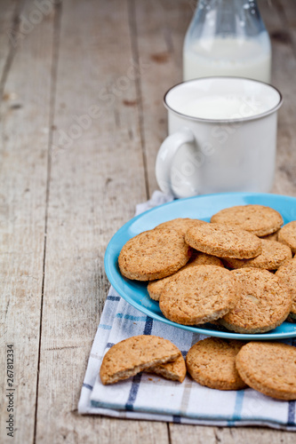 Fresh baked oat cookies on blue ceramic plate on linen napkin and cup of milk on rustic wooden table background.