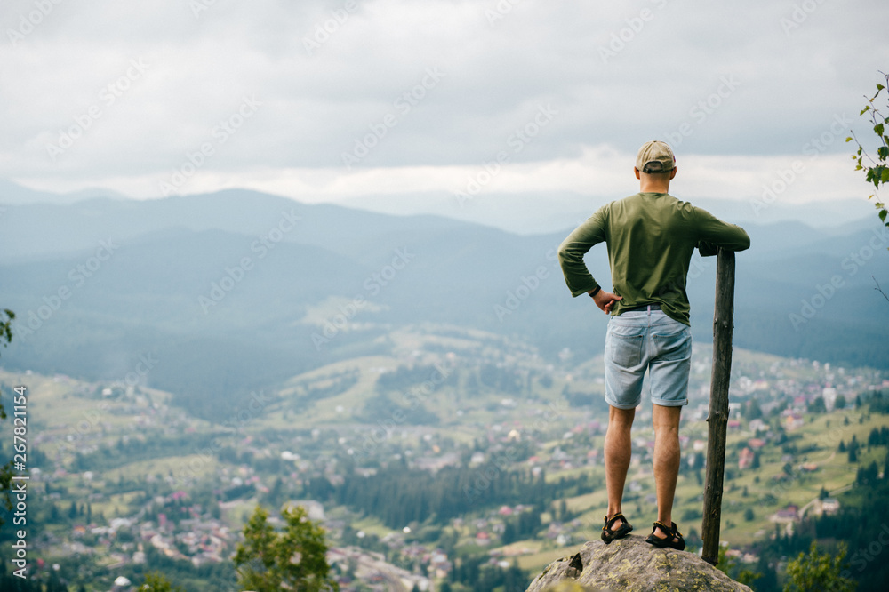 Lifestyle summer portrait from behind of successful man with wooden stick standing on top of mountaing with beautiful landscape in front. Male traveler enjoying nature view from highest peak at hill.