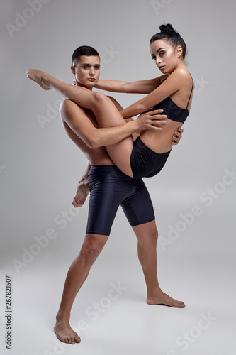 The couple of a young modern ballet dancers in black suits are posing over a gray studio background.