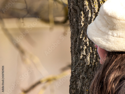 Obraz na plátně A young woman with gray eyes, a white hat and warm clothes embracing a tree trun