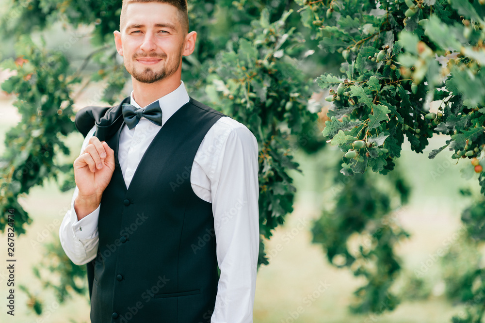 Lifestyle portrait of happy groom posing for camera outdoor at nature with oak tree on background. Cheerful fiance with smiling face in jakcet, bowtie and white shirt portrait before wedding ceremony. Lifestyle portrait of happy groom posing for camera outdoor at nature with oak tree on background. Cheerful fiance with smiling face in jakcet, bowtie and white shirt portrait before wedding ceremony.