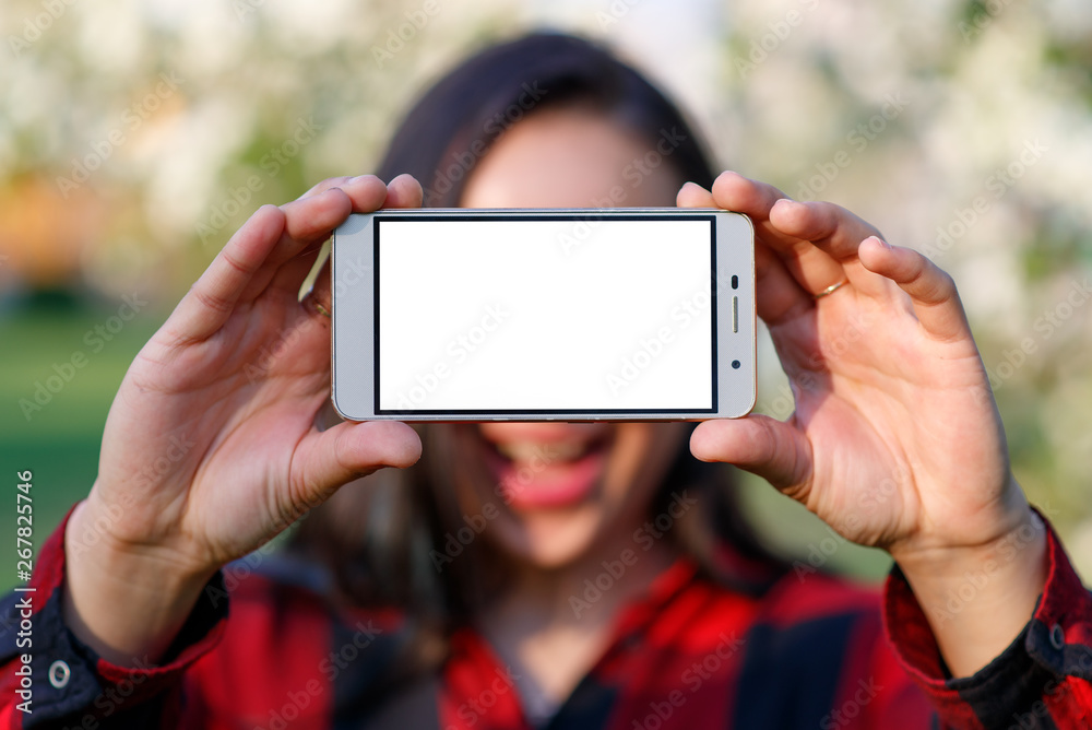 A young woman holds a smartphone with an empty white screen in front of her with two hands. With a place to mine space. Outdoors, in summer on a Sunny day