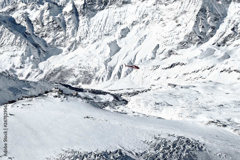 Aerial shot of a red rescue service helicopter flying in the winter in the Swiss Alps against the mountains above the cracks in the glacier. you can see a large number of rocks and snow   