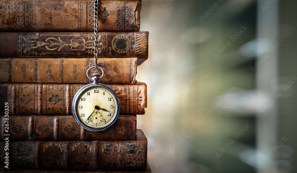 Vintage clock hanging on a chain on the background of old books. Old watch  as a symbol of passing time. Concept on the theme of history, nostalgia,  old age. Retro style. Stock
