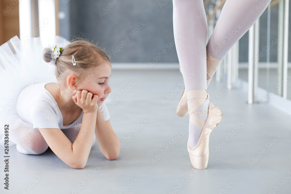 Little ballerina is looking at legs of professional ballet dancer in pointe shoes. Cute kid girl dreaming. Concept of classical dance school, practicing for children.