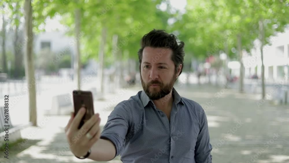 Handsome man taking selfie with smartphone outdoor. Smiling bearded man holding cell phone and photographing on street. Digital device concept Handsome man taking selfie with smartphone outdoor. Smiling bearded man holding cell phone and photographing on street. Digital device concept