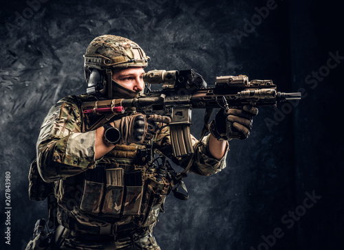 Close-up studio photo against a dark textured wall. The elite unit, special forces soldier in camouflage uniform holding an assault rifle with a laser sight and aims at the target