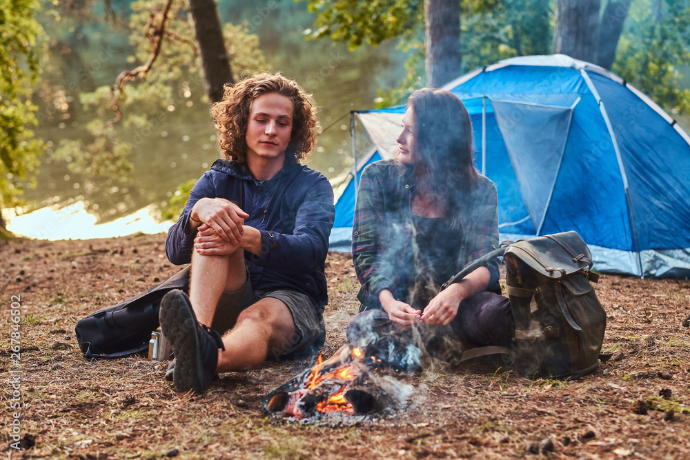 Young happy students are chilling and talking near bonfire in the green forest. There are tent and lake nearby. Young happy students are chilling and talking near bonfire in the green forest. There are tent and lake nearby.