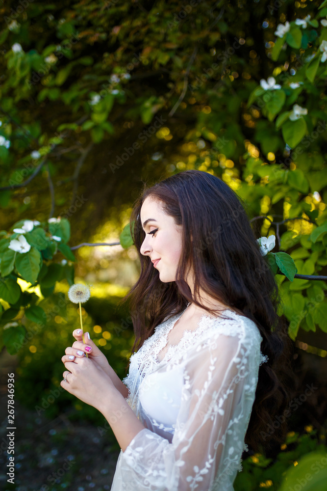 young girl in white dress with long dark wavy hair holding a dandelion in the garden