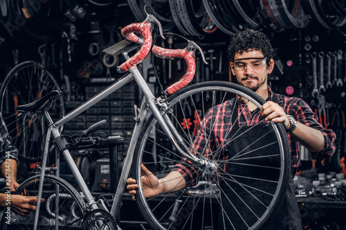 Hardworking repairmans are fixing bicycle at their busy workshop. There are a lot of tools at background.