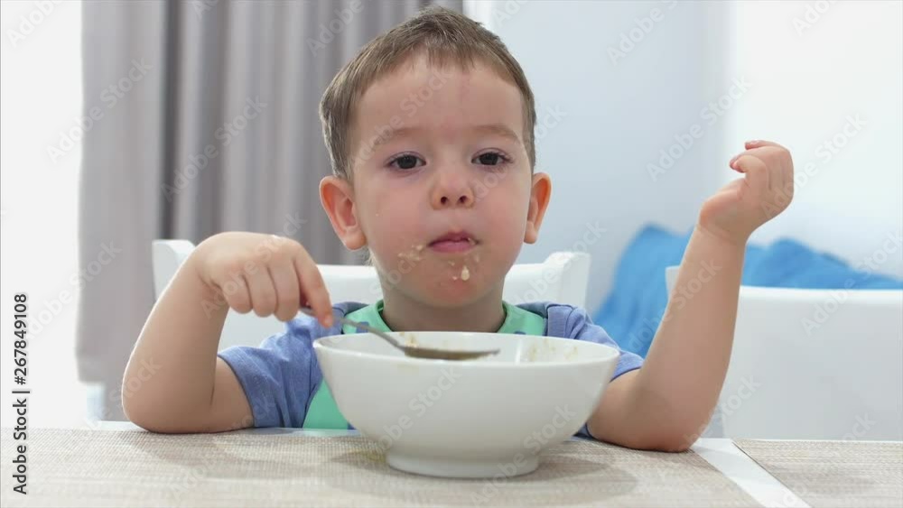 Small Cute Child is Sitting at a Table and eat his own oatmeal, the baby eats willingly. Concept Happy Childhood. Small Cute Child is Sitting at a Table and eat his own oatmeal, the baby eats willingly. Concept Happy Childhood.