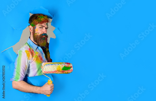 Repair. Painter with painting roller through hole in paper. Professional painter in dirty shirt making hole in paper wall. Serious painter man. Handsome bearded worker with paint roller. Copy space. photo