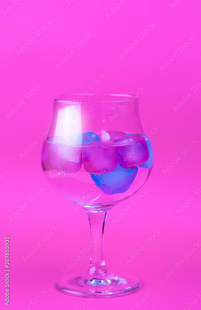A cold drink with colorful pink and blue ice in a wine glass on a bright pink background. Summer concept horisontal image