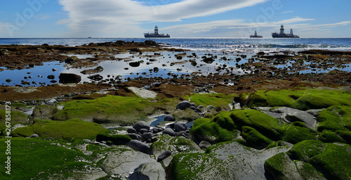 Coast at low tide in foreground, calm sea, blue sky with clouds and oil rigs in the bay 