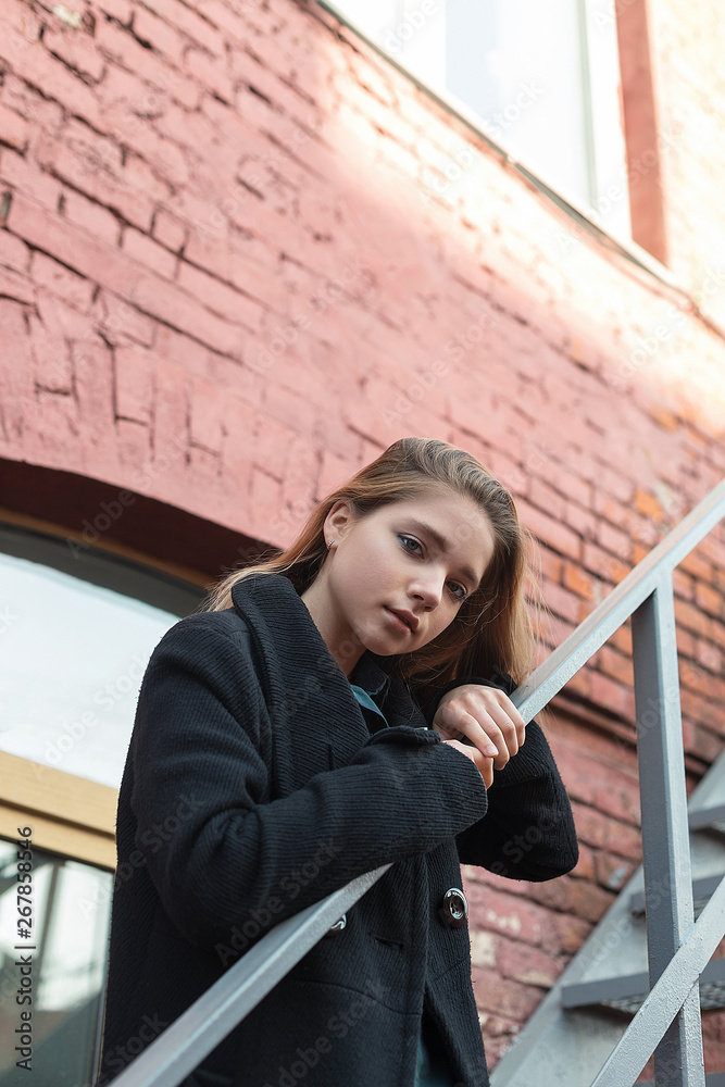Young girl in black coat standing on stairs with brick wall on background. Concept of loneliness.