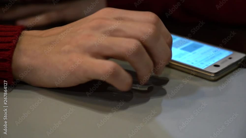 Close-up of a young guy's hand with scars and veins in a red sweater and a white phone with an included screen. The man is nervous, knocking his fingers on the table and waiting for download. Close-up of a young guy's hand with scars and veins in a red sweater and a white phone with an included screen. The man is nervous, knocking his fingers on the table and waiting for download.