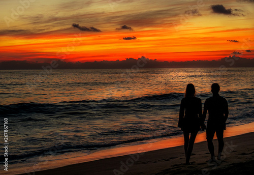 Man and woman couple in love holding hands during red sunset at the beach with waves at the ocean in Mexico