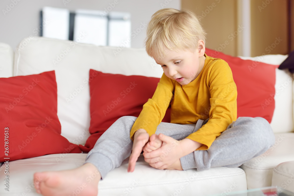 Little boy examines the sore leg sitting on the couch indoors. Trauma ...
