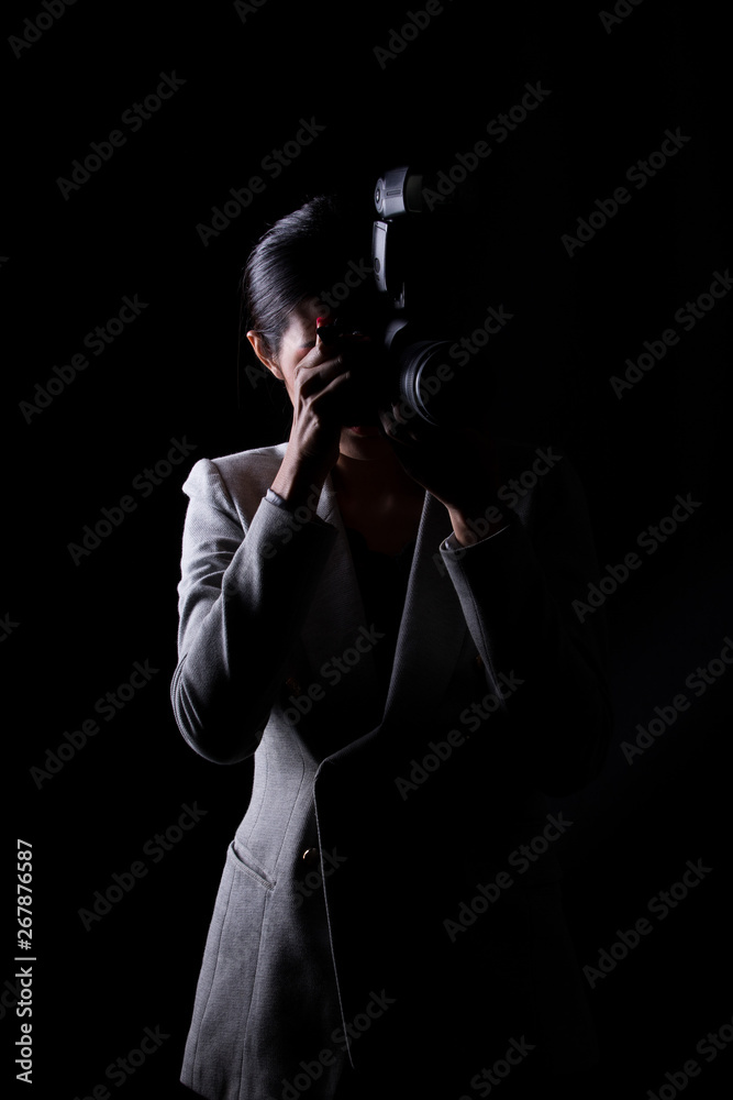 Asian Woman Photographer hold camera with external flash point to shoot subject, wear gray suit. studio lighting black background isolated low key exposure, reporter journalist take photo celebrity Asian Woman Photographer hold camera with external flash point to shoot subject, wear gray suit. studio lighting black background isolated low key exposure, reporter journalist take photo celebrity