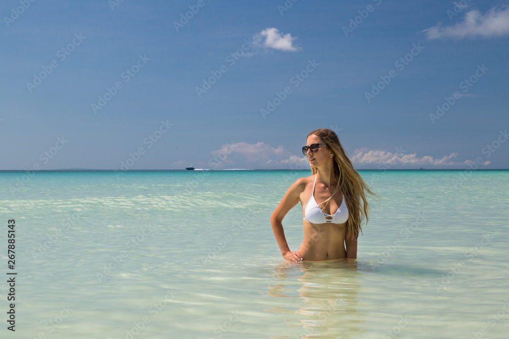 Sexy tanned girl in white swimsuit posing on the sea. Beautiful model sunbathes and rests on ocean shore. Concept vacation, travel, swimwear