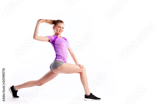 A young woman coach in a sporty short top and gym leggings makes lunges by the feet forward, hands are held out to the side on a white isolated background in studio