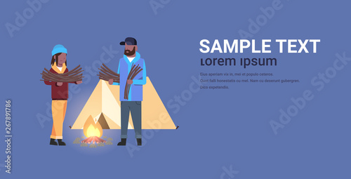 couple tourists hikers holding firewood african american man woman organizing fire camp with canvas tent and campfire hiking camping concept horizontal full length copy space flat