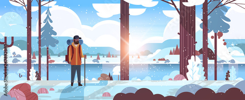 tourist hiker with backpack african american man traveler holding stick standing in winter forest hiking concept sunrise snowfall landscape nature river mountains background horizontal full length