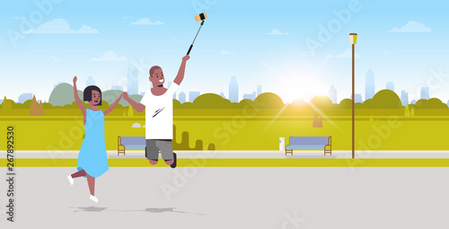 young couple using selfie stick taking photo on smartphone camera african american man woman jumping having fun city urban park sunrise landscape background full length horizontal