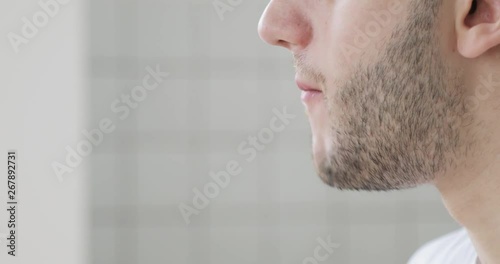 Portrait of young bearded hungry man is eating tasty burger at home at light background, mouth closeup, side view. photo