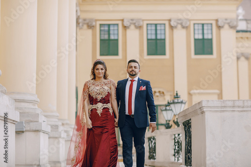 Beautiful woman in a long red dress walks around the city with her husband in a blue suit and with a beard